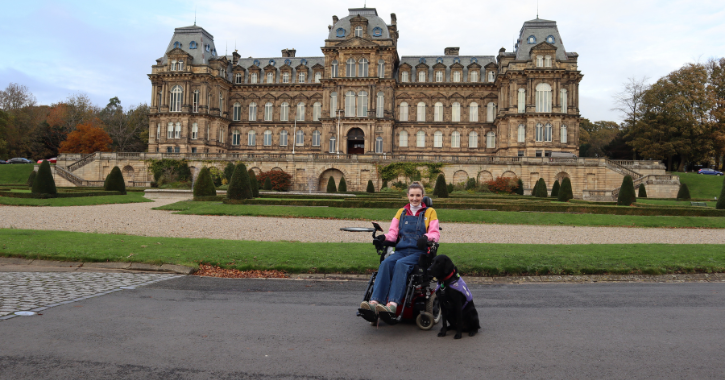 woman sat in wheelchair with service dog by her side with The Bowes Museum in the background.